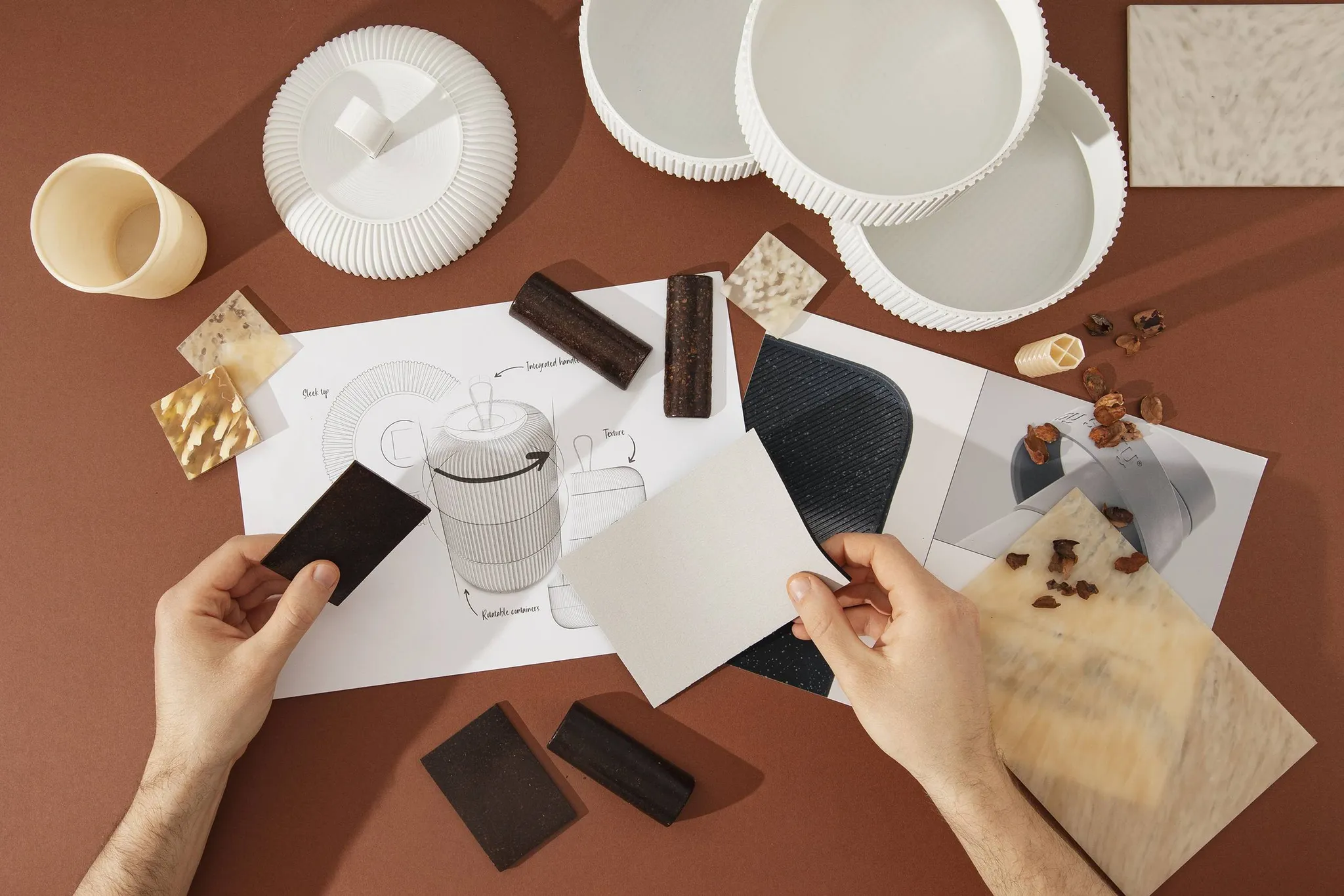 Two hands shown holding material samples over a series of sketches, drawings, material samples and 3D models