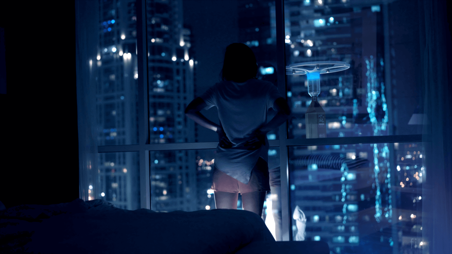 A woman stands inside her skyscraper apartment, with large windows. Other skyscrapers can be seen through her window. A drone carrying a package is hovering outside her window