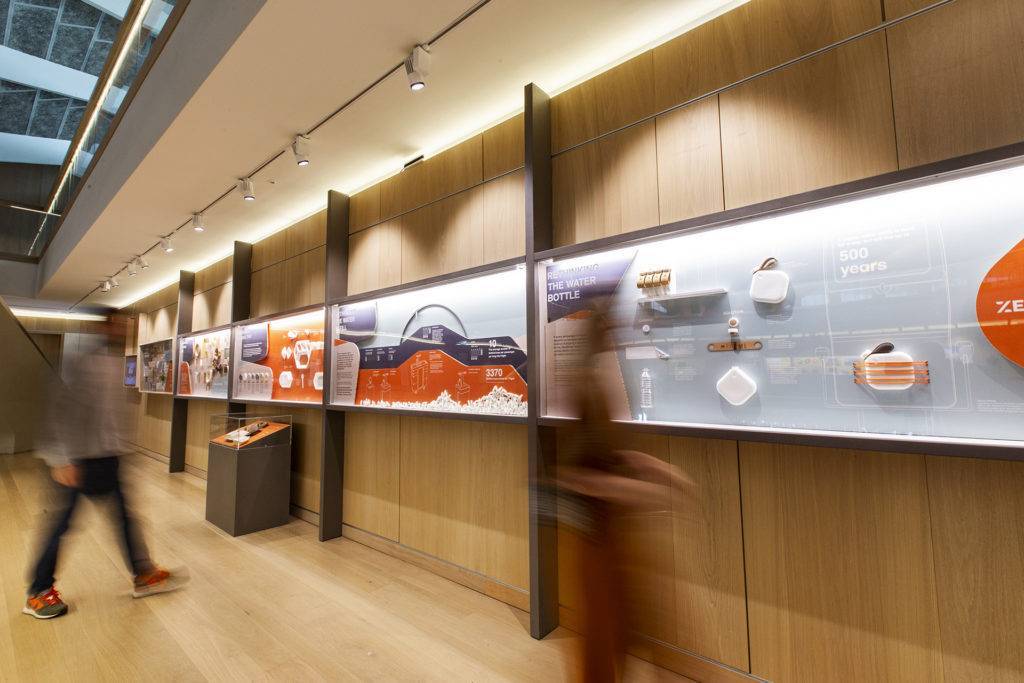 Exhibition view of Get Onboard at the Design Museum, with display cases on the wall. Two blurred people can be seen looking at the displays