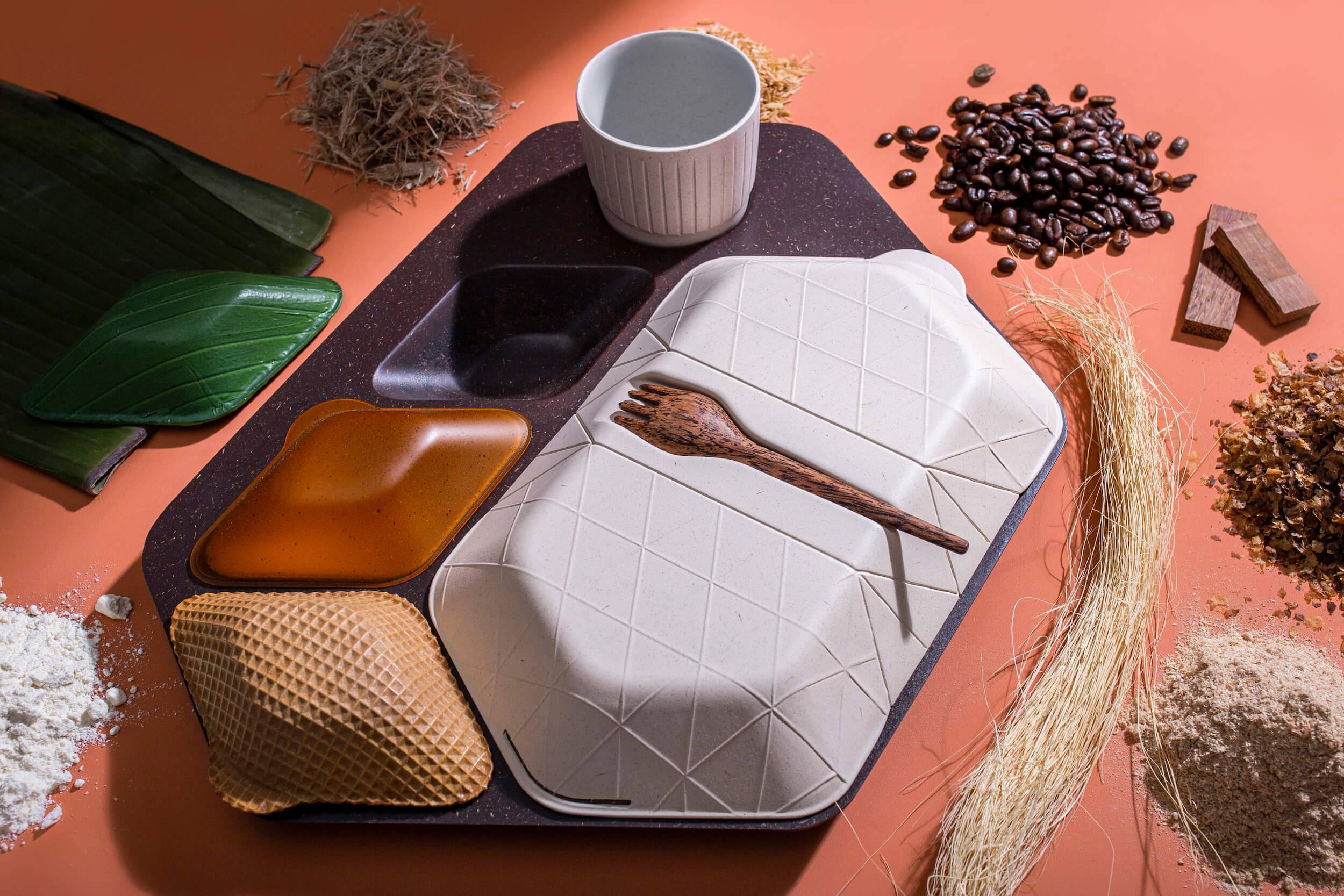 An airline mealtray made of sustainable materials, including wafer biscuit, algae, coffee grounds and coconut wood, shown alongside the raw materials that it is composed of