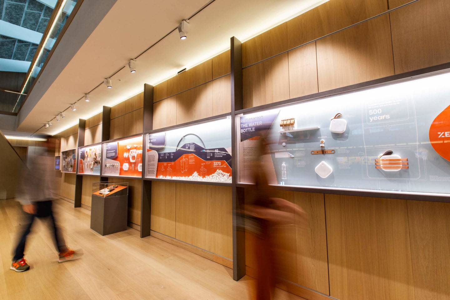 Exhibition view of Get Onboard at the Design Museum, with display cases on the wall. Two blurred people can be seen looking at the displays