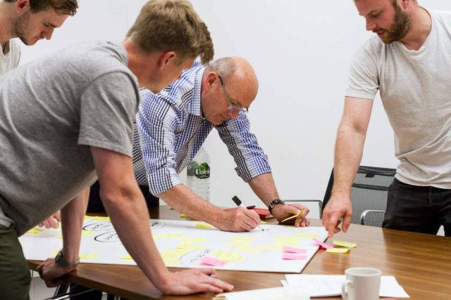 Four people leaning over a table, one of them drawing on a large sheet of paper