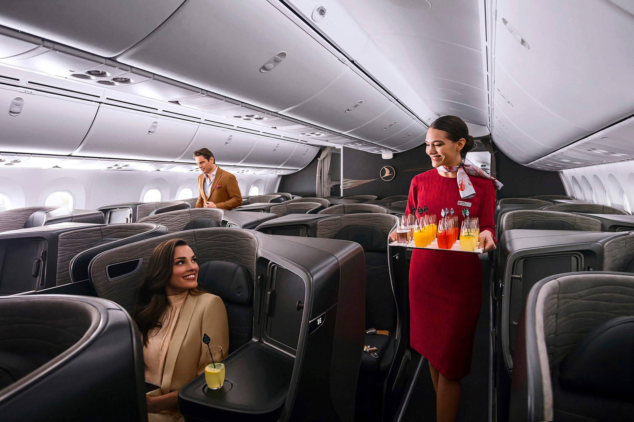 Cabin crew walking down a business class cabin holding a tray filled with drinks, a seated passenger smiles at her. Another passenger is seen standing in the back