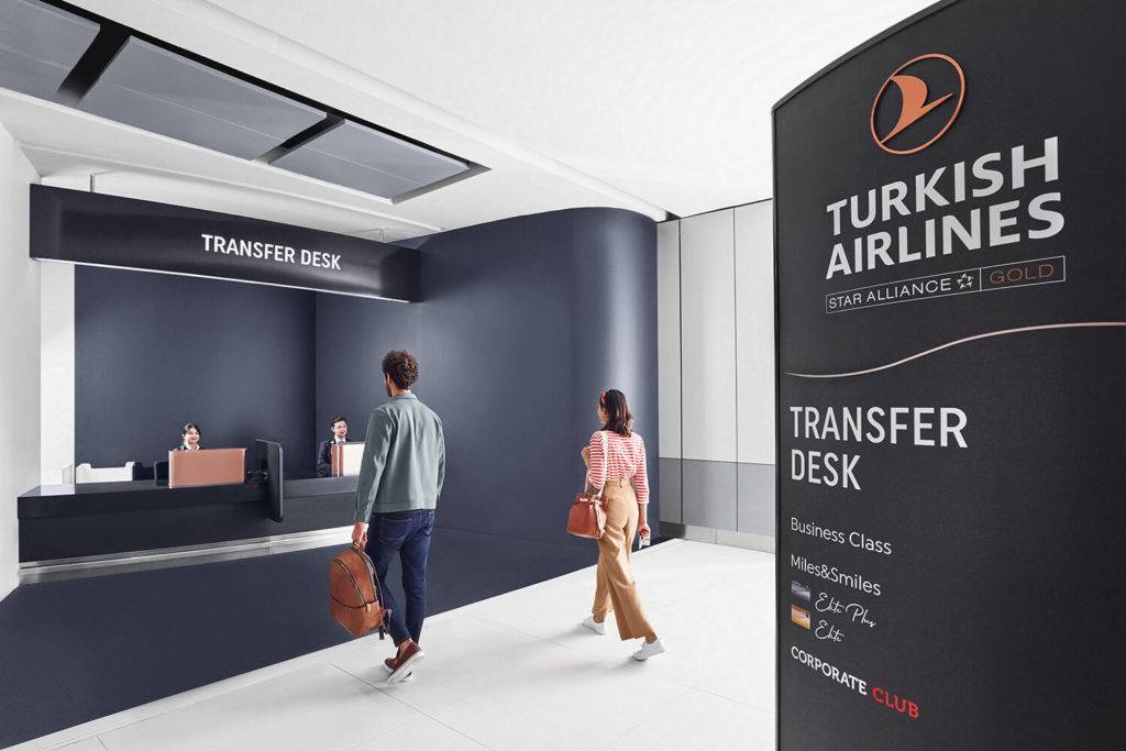 Turkish Airlines transfer desk. Two passengers are seen walking towards a transfer desk. Wayfinding signage is seen on the right