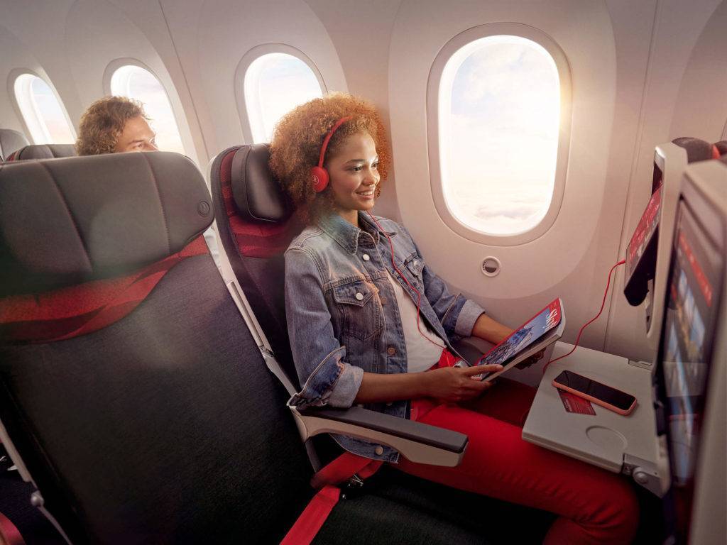 A woman sitting in an economy class seat, holding a magazine and watching the screen from the IFE system