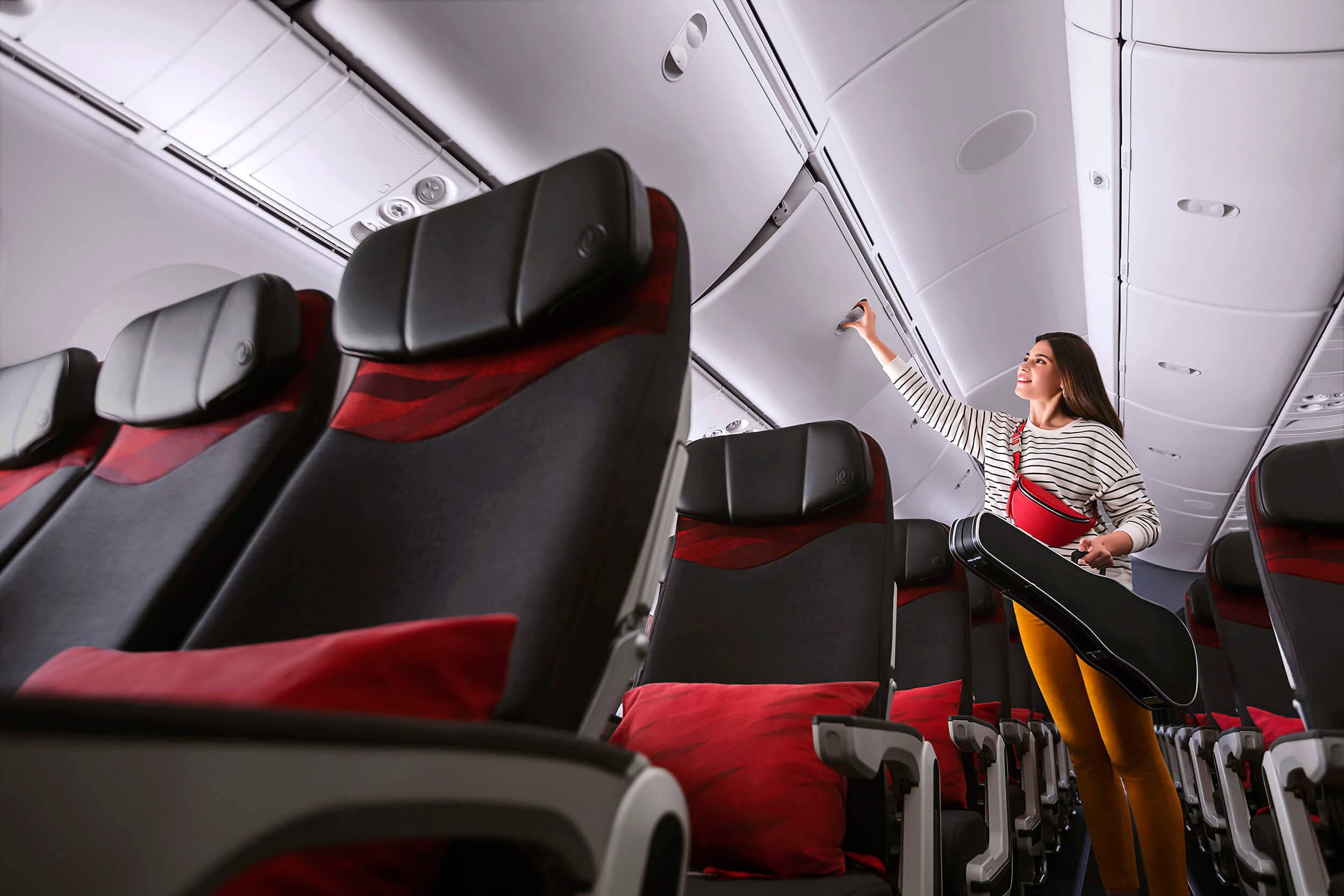 A woman reaches to open an overhead luggage bin to store a violin in economy class