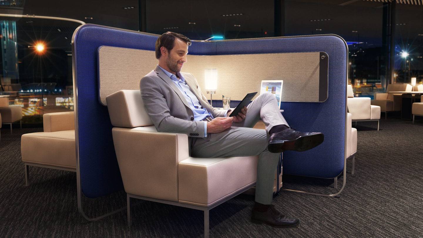 A man seated in a lounge chair surrounded by upholstered privacy panels, with a small table lamp in an airport lounge