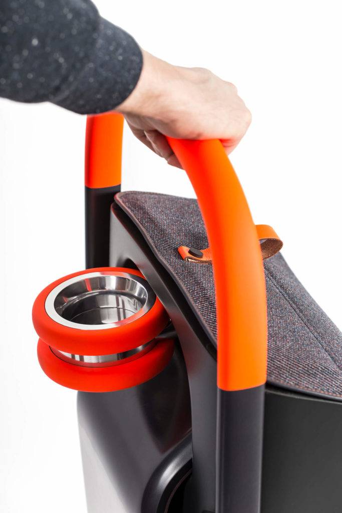 A hand holding the ergonomic handle of a personal mobility scooter. A storage bag can be seen, with integrated cup holder