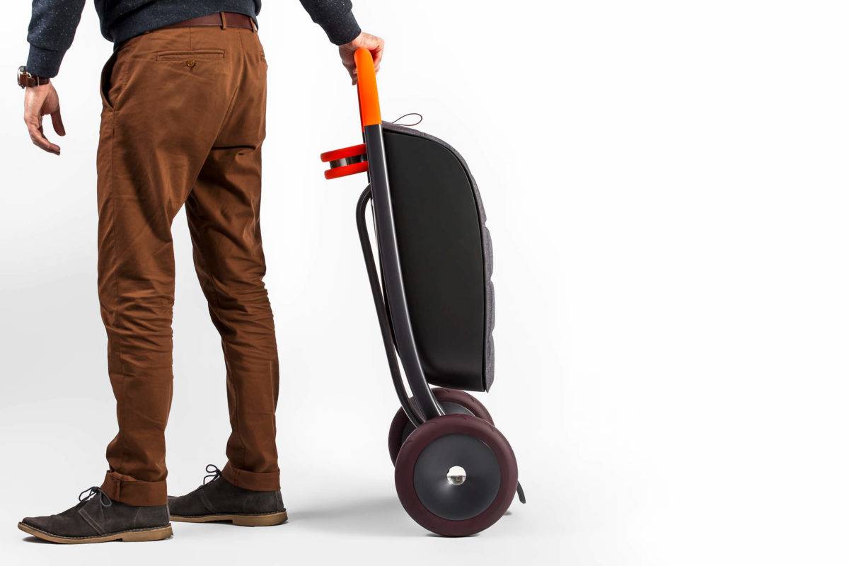 A person holding a folded personal scooter, which features a large storage bag in the front and a cup holder by the handles