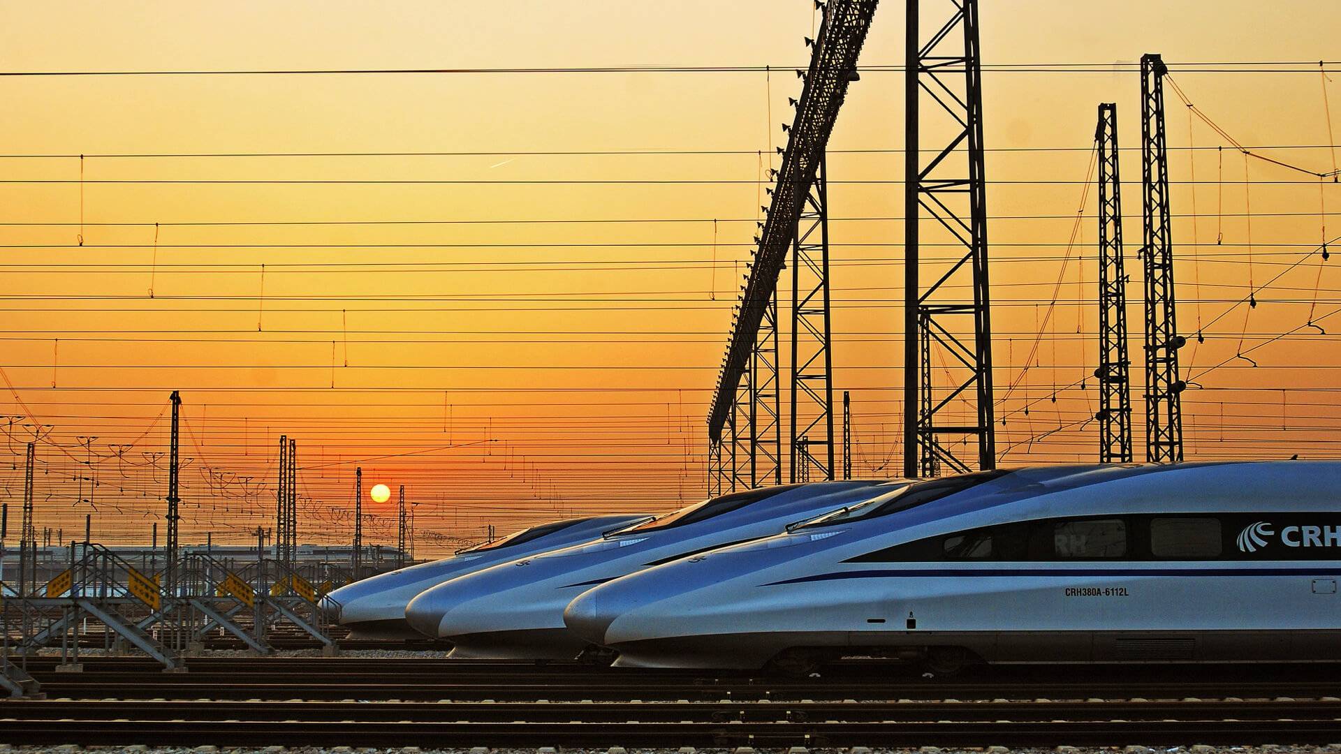 The streamlined noses of three high speed trains stopped on tracks, with a sunset in the background