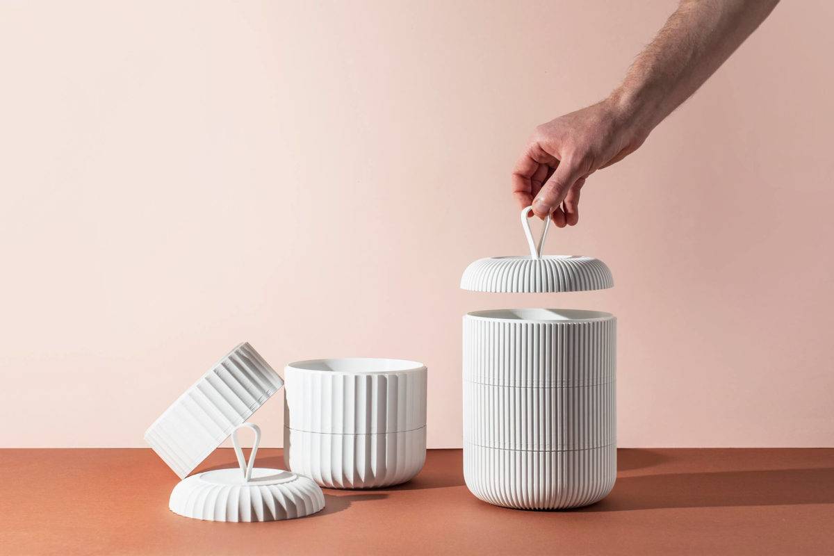 A hand lifting the lid of a white 3D printed model of the bento box style stacked food containers. A second, partly unstacked model sits on the left. The products are laid out on a terracotta surface against a light pink background