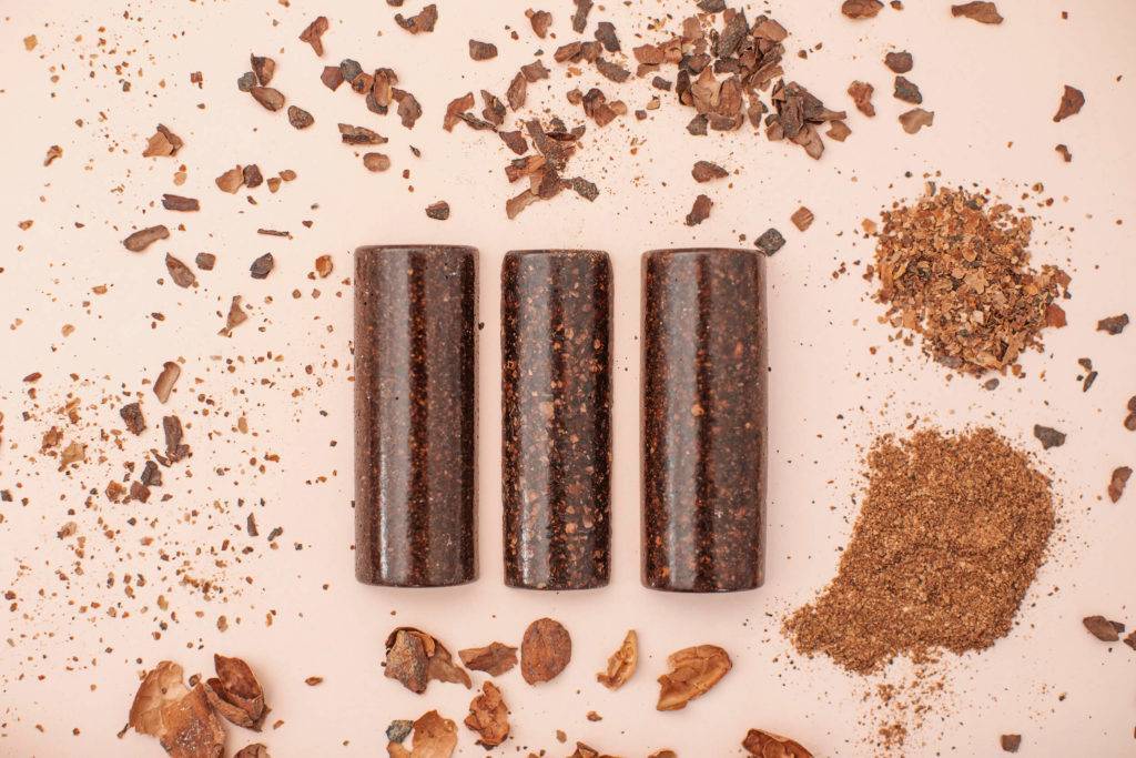 Three samples of a natural material made from cacao, surrounded by sprinkles of the raw material, set out on a light pink background