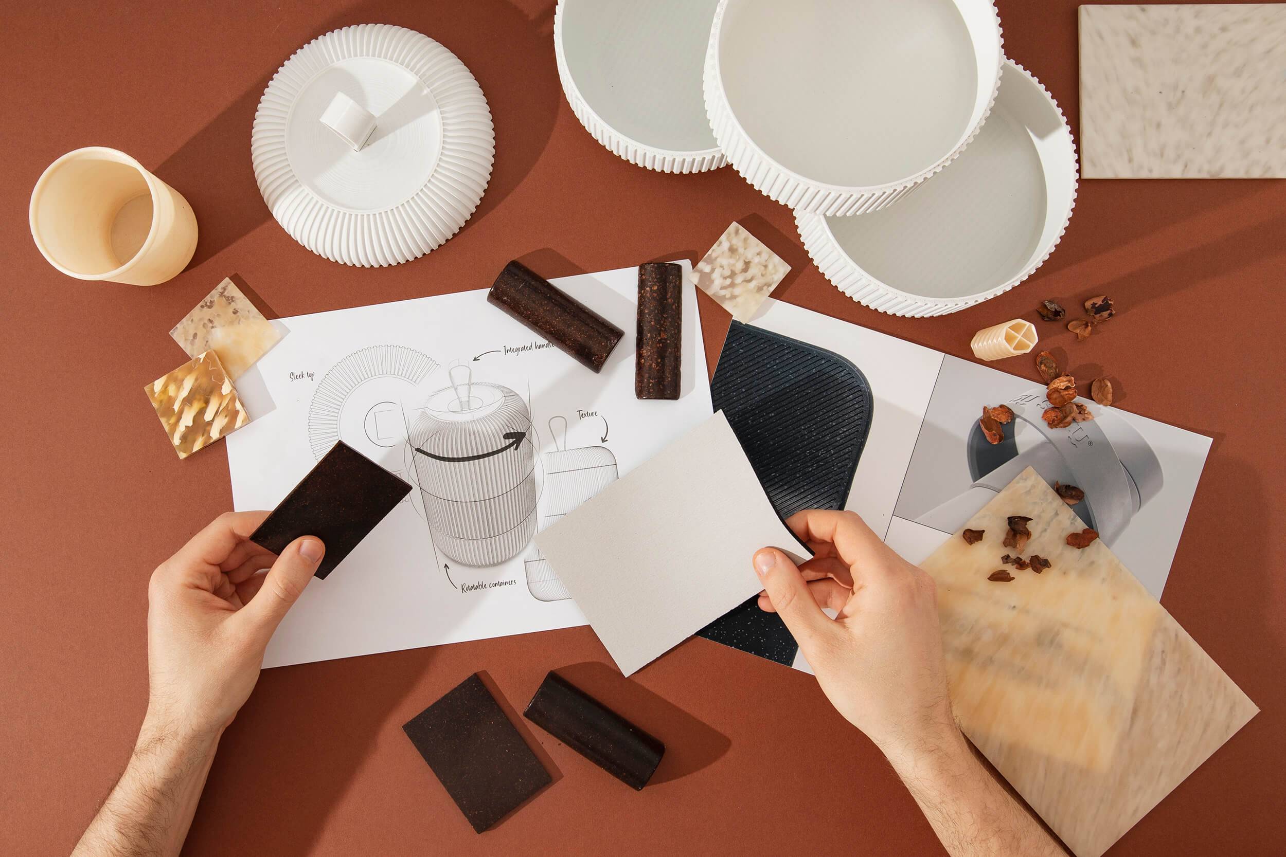 Two hands shown holding material samples over a series of sketches, drawings, material samples and 3D models