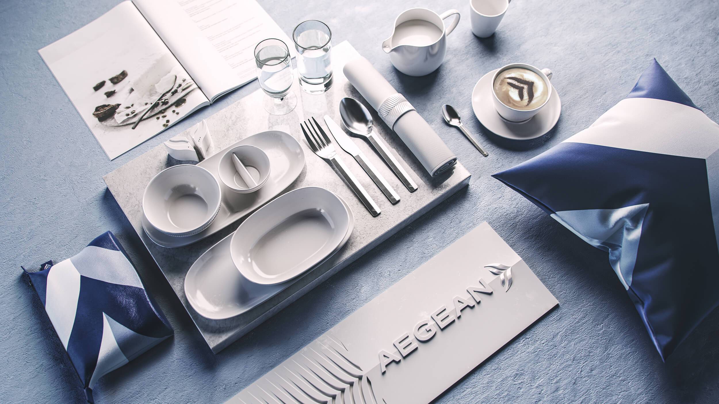 Overhead shot of the Aegean Airlines service items, including a branded amenity kit, branded cushion and meal items. An Aegean 3D brand panel features beneath, and the food menu above