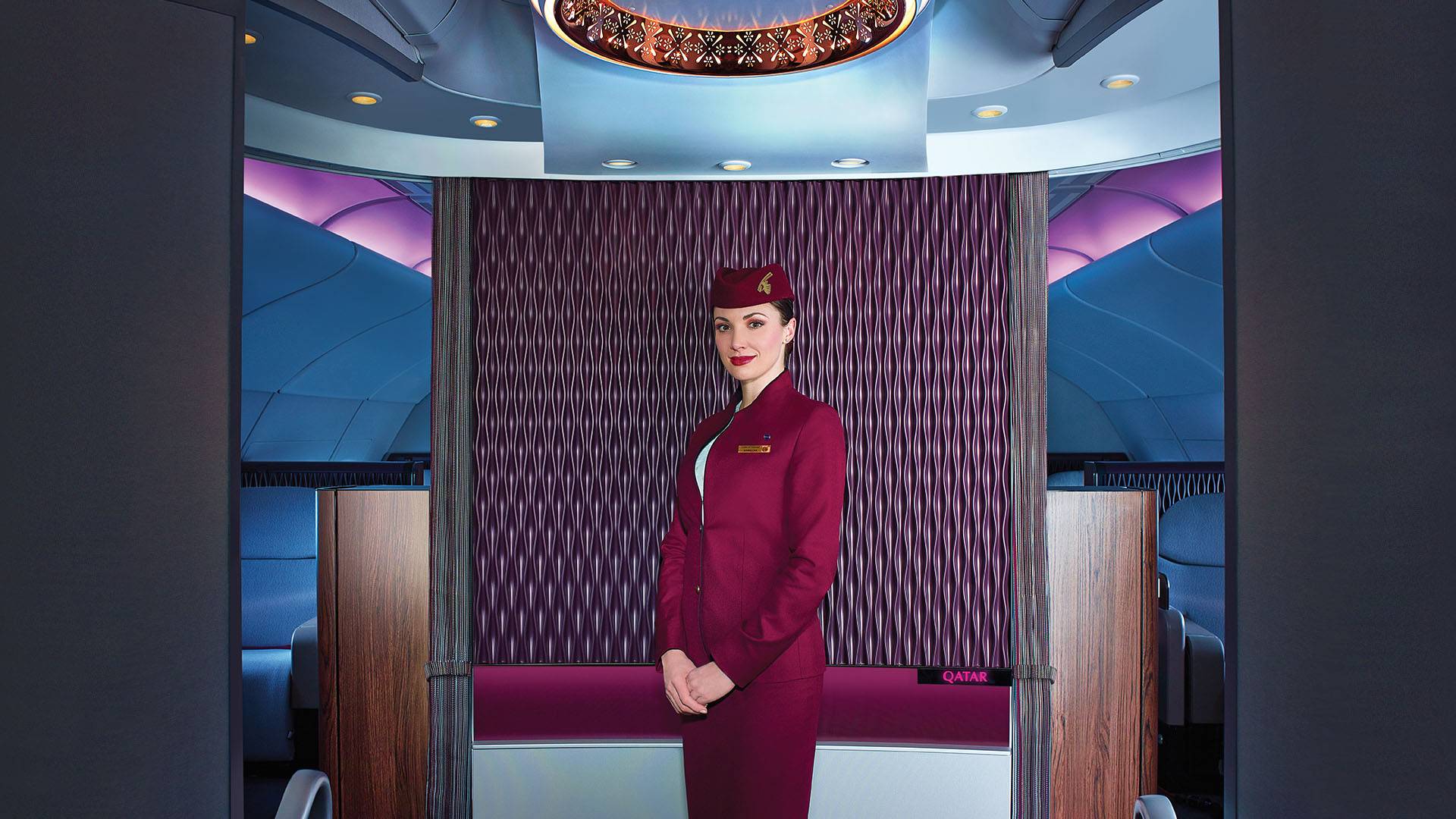 A cabin crew stands in the entrance area to Qatar Airways A380 First Class cabin, with a statement light and feature brand panel