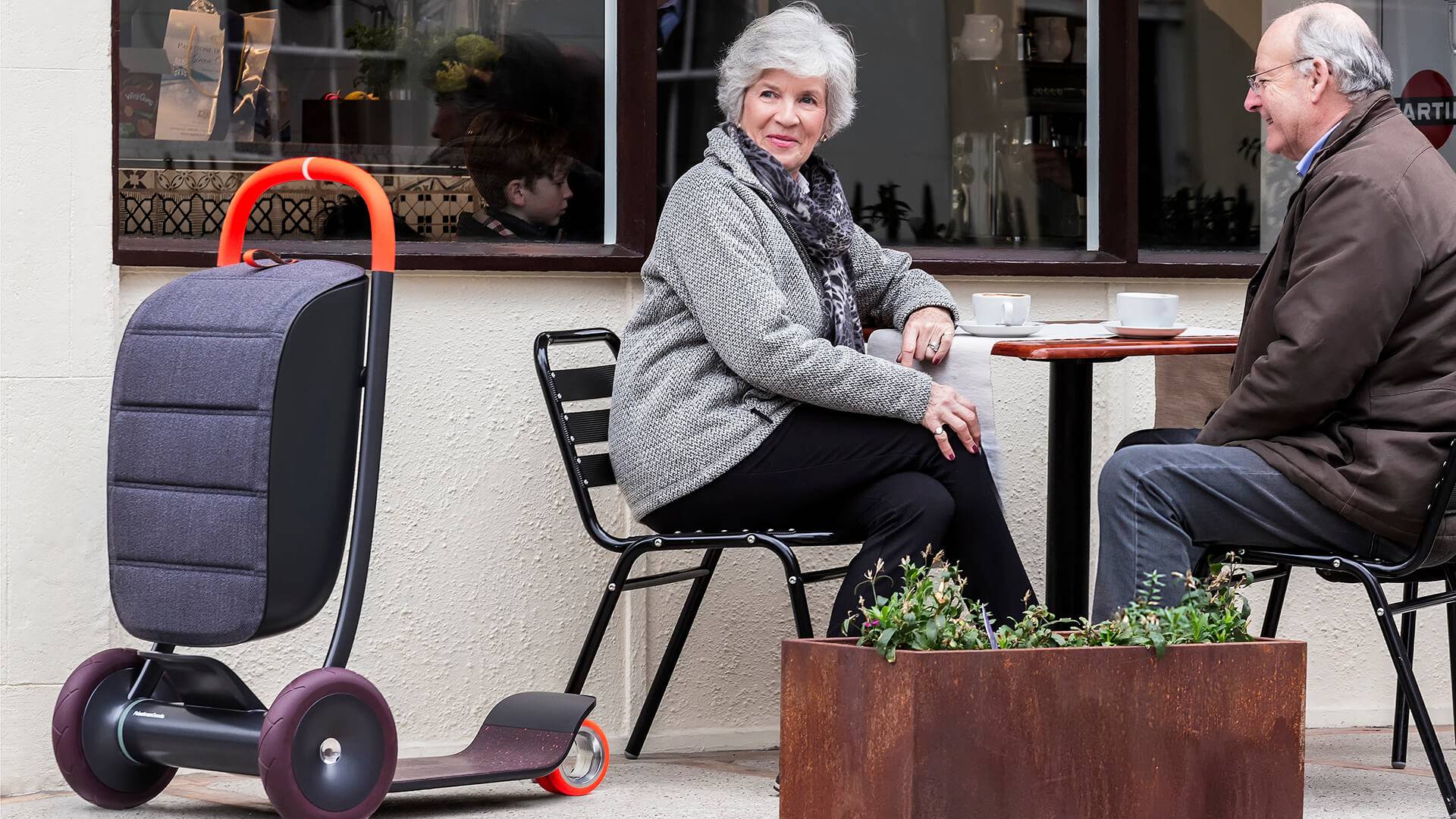 An older couple sit at an outdoor table in a cafe, with a three-wheeled personal scooter next to the woman