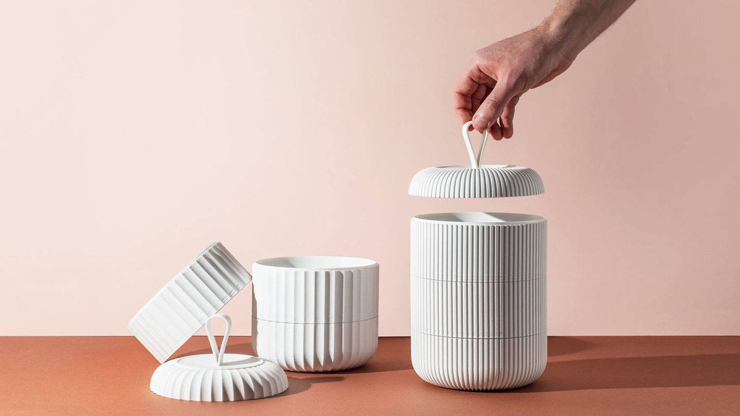 A hand lifting the lid of a white 3D printed model of the bento box style stacked food containers. A second, partly unstacked model sits on the left. The products are laid out on a terracotta surface against a light pink background