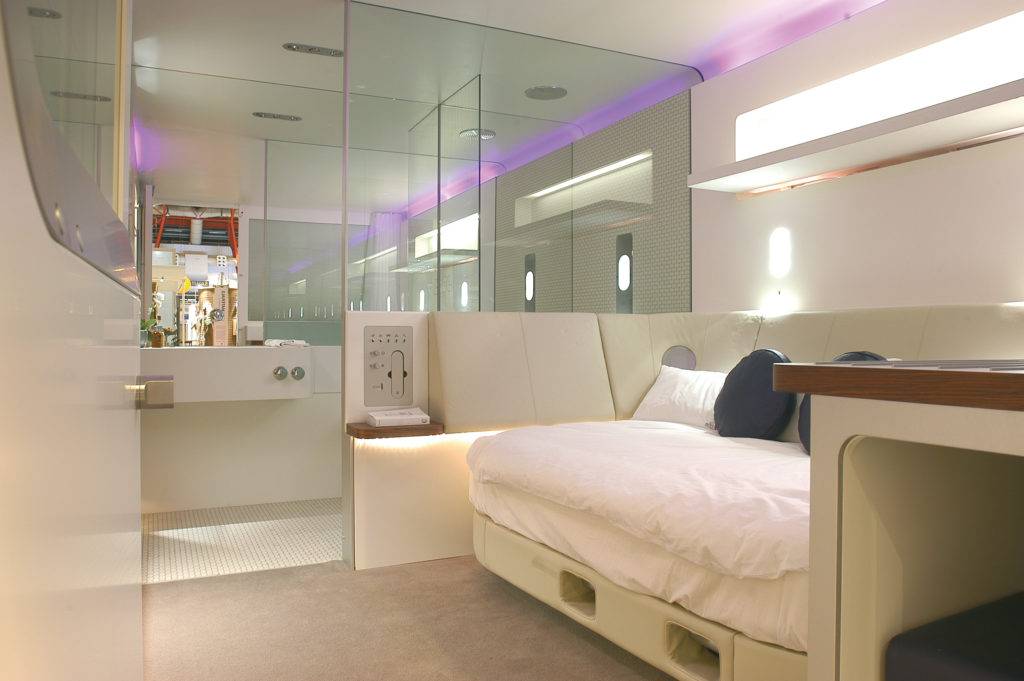 A compact hotel room with a sofa doubling as a bed, which has a padded surround, a glass partition leads to a bathroom