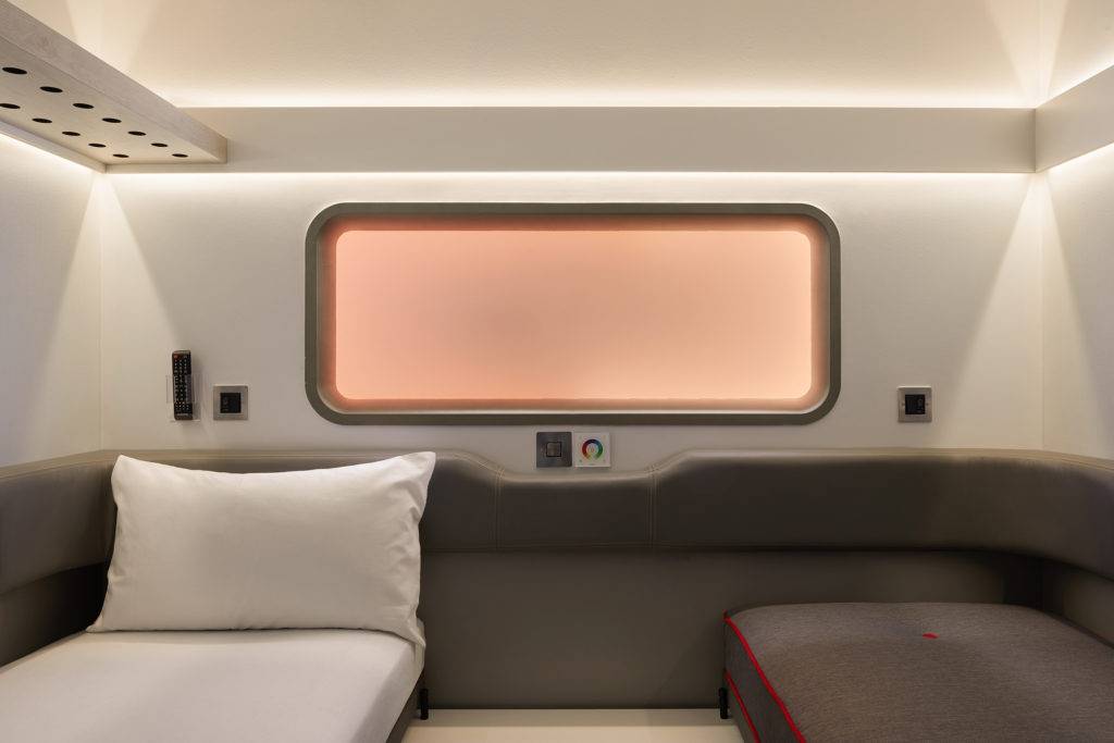 A compact hotel room with a bed on one side, a sofa on the other, a padded surround on the wall, recessed lighting in the ceiling, a storage shelf above the bed and an orange frosted window